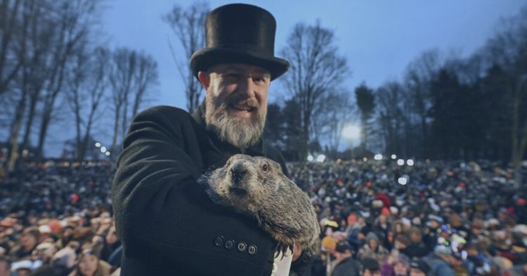 Recruiting Trends 2025: A man wearing a black coat and top hat holds a groundhog in his arms, smiling at the camera. Behind them, a large crowd gathers outdoors in a wooded area during twilight, with trees and soft lighting visible in the background.