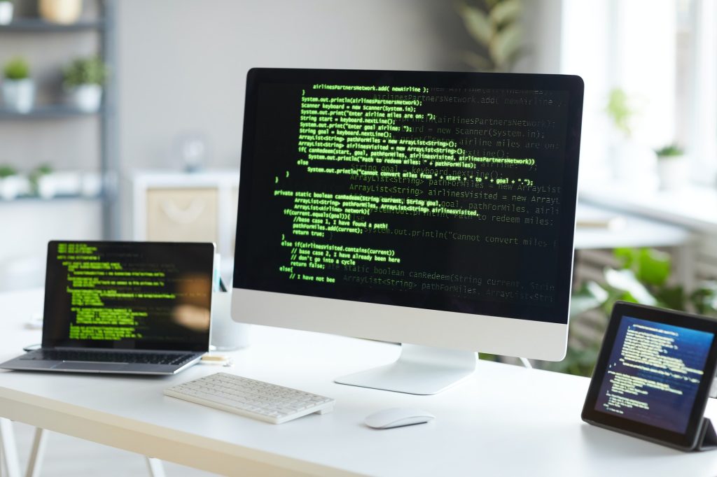 A workspace with a desktop monitor, laptop, and tablet displaying green code on black screens, set in a modern office.