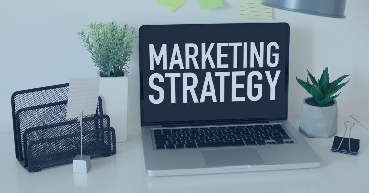 Recruitment Marketing Strategy: A workspace featuring an open laptop with the words "MARKETING STRATEGY" displayed on the screen. The desk is decorated with potted plants, a file organizer, and office supplies, conveying a professional and organized setup.