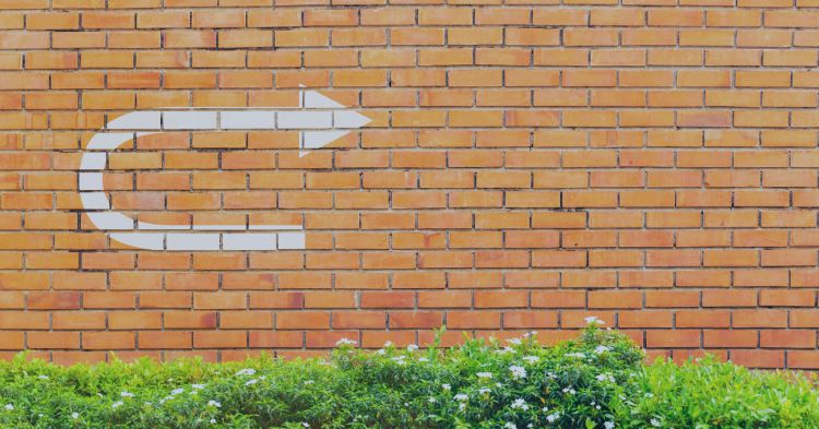 Staffing Firm Turnaround Strategies: A brick wall with a white painted U-turn arrow, positioned above a line of green plants with small white flowers, suggesting a change in direction or a shift in perspective.