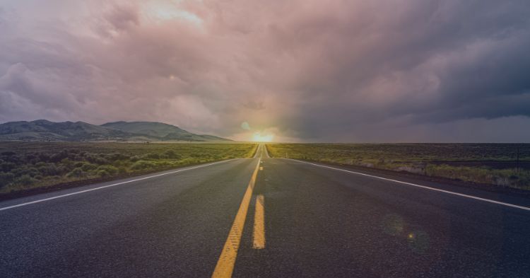 Strategic Staffing: A straight, two-lane road stretches into the distance, framed by green fields and low hills under a dramatic cloudy sky. The setting sun casts a warm glow on the horizon, creating a sense of depth and calm.