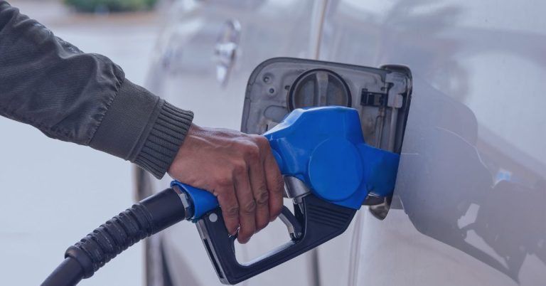 JobDiva Technology: A person refueling a silver vehicle with a blue gas pump nozzle at a gas station. The person's hand grips the nozzle, and the fuel door is open.