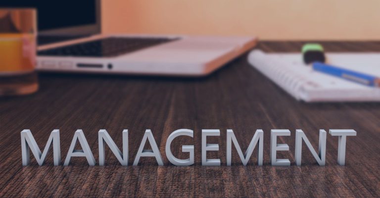 Staffing Management: The word 'MANAGEMENT' in bold, three-dimensional letters on a wooden desk, with a blurred background featuring a laptop, notebook, pen, and a highlighter, symbolizing organization and leadership.